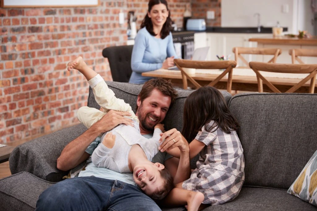 A father and two children play on the sofa while the mother sits at a table in the background and works on a laptop