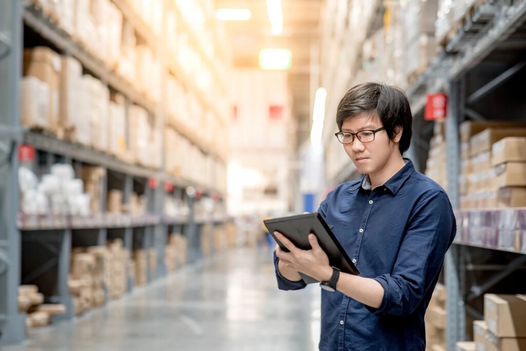 Man with dark blue shirt and glasses reading a tablet while standing inside a warehouse