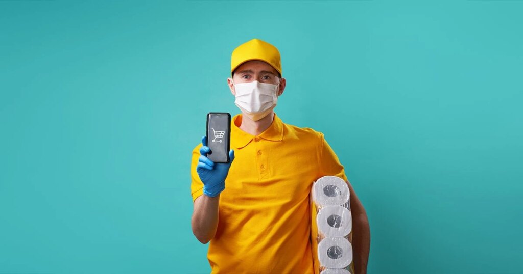 Courier dressed in yellow with gloves and mask, holding toilet paper and phone