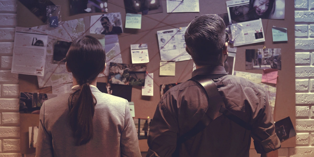 Two detectives, one male and one female, studying a bulletin board of crime scene case evidence