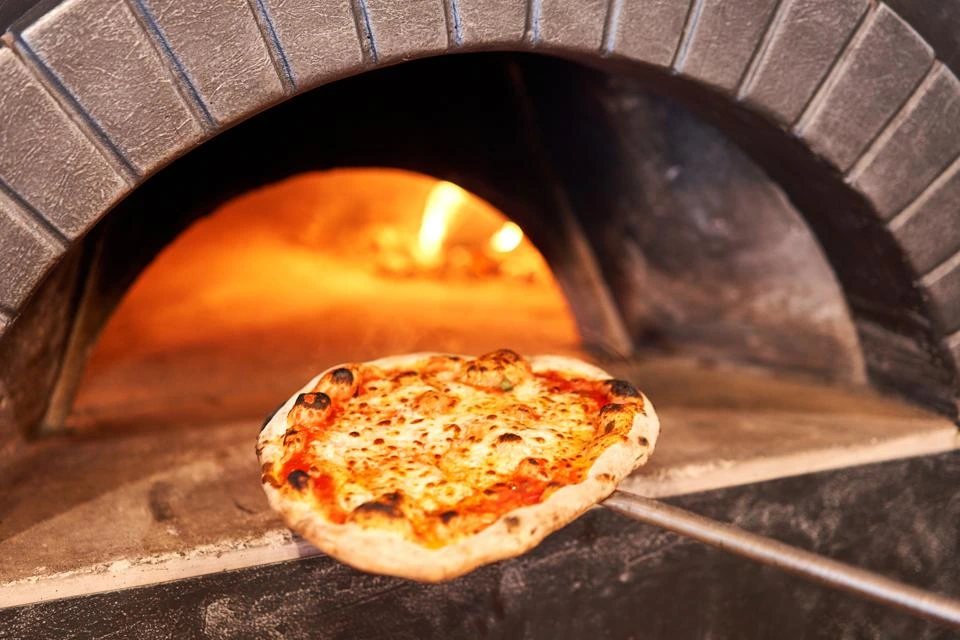 Small artisan pizza on wooden paddle being placed in traditional pizza oven